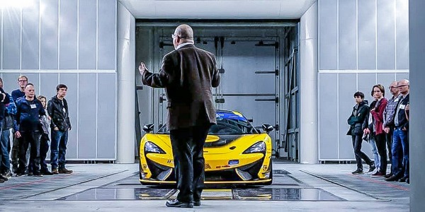 Man stands brightly lit in front of a yellow sports car, speaking to a crowd of individuals to the right and left.