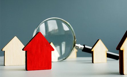 A single red wooden house token stands amongst natural wood house tokens and a magnifying glass