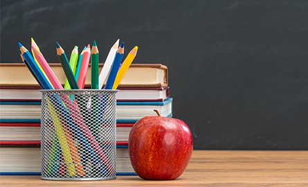 An apple sitting on a desk beside pencil crayons and in front of a stack of books