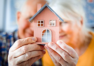 Two seniors hold a wooden house in front of their faces