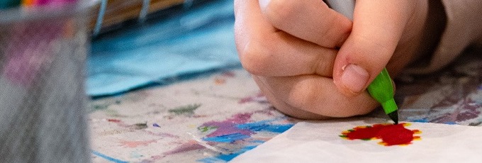 A girls hand using a green marker to outline a red heart in the corner of a white square piece of paper. A silver metal container of markers out of focus on the side.