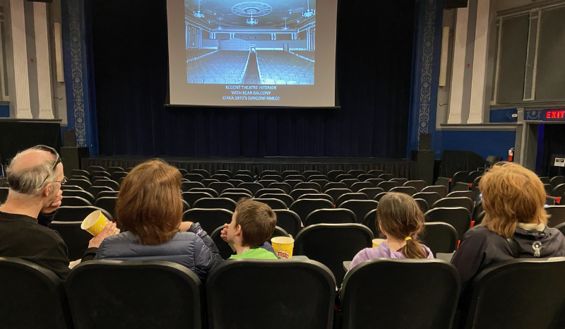 Family of five in theatre seats facing large screen and stage eating popcorn.